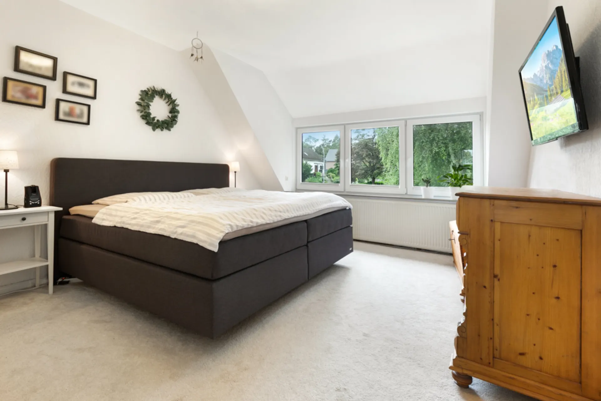 Helles Schlafzimmer mit großem Bett, Holzkommode, Fenstern und Wandfernseher.