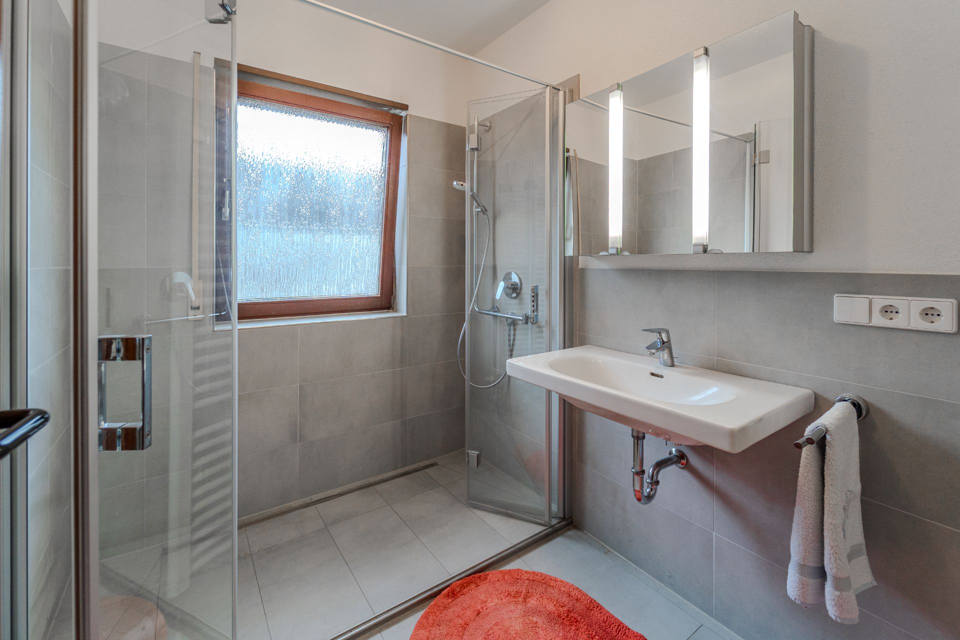 Modernes Badezimmer mit Dusche, Waschbecken, Spiegel, Fenster und rotem Teppich.
