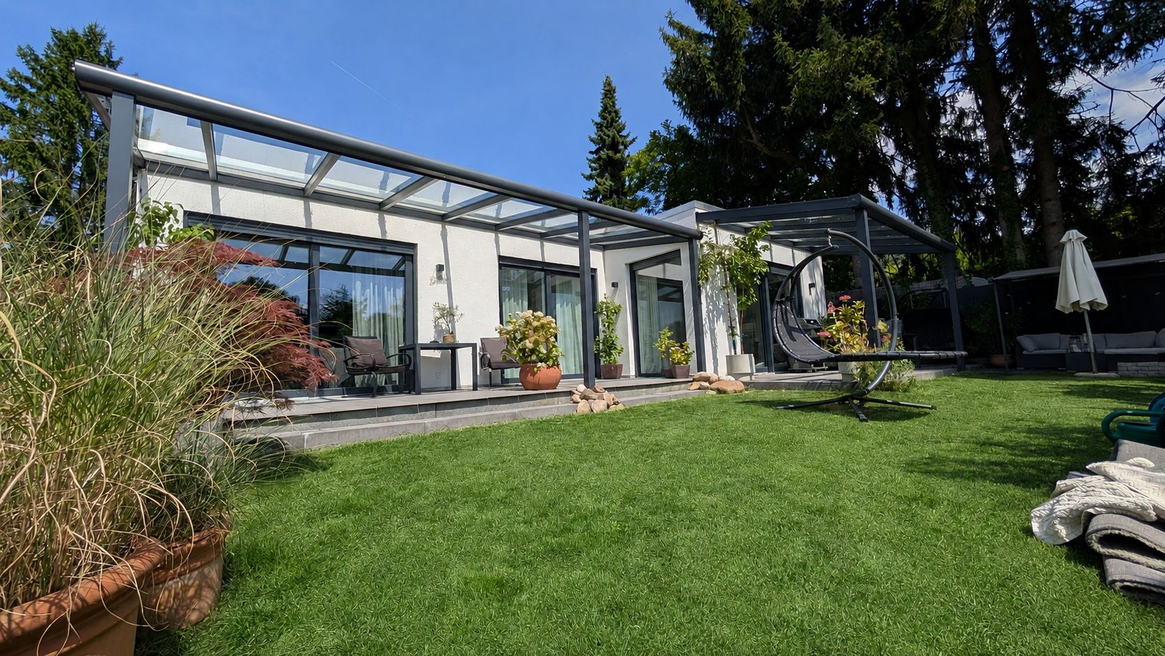 Modernes Wohnhaus mit großer Glasüberdachung, Terrasse und Garten mit Rasen und Pflanzen.