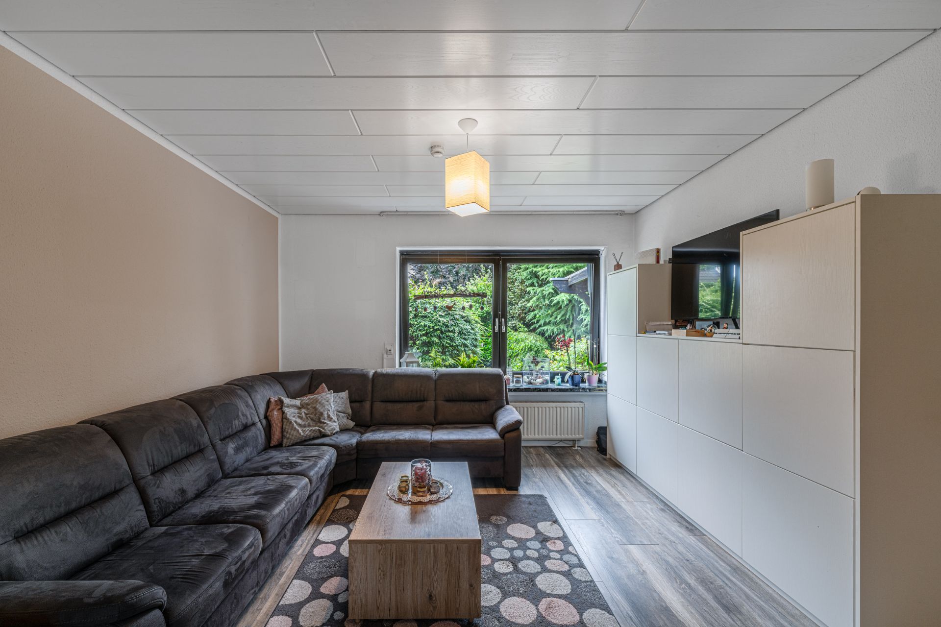 Wohnzimmer mit großer Eckcouch, Couchtisch, TV und Fenster mit Blick ins Grüne.