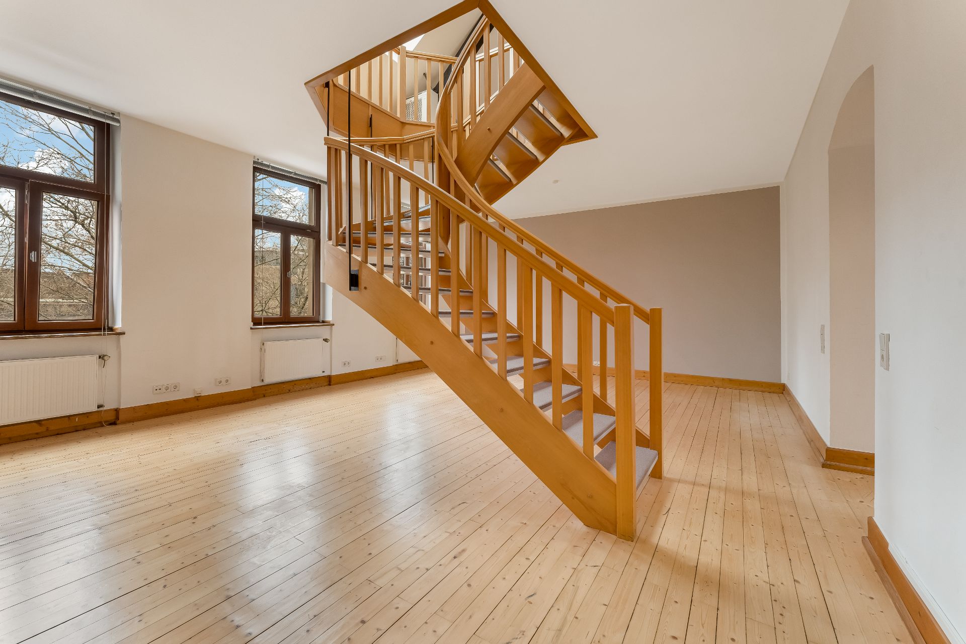 Helles Zimmer mit Holztreppe in der Mitte und großen Fenstern zur linken Seite.