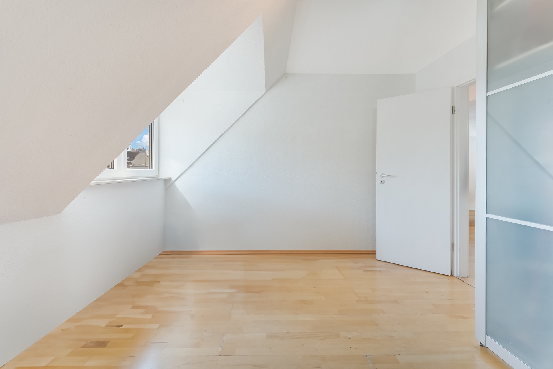 Helles Zimmer mit Dachschräge, Holzfußboden, offenem Fenster und geöffneter weißer Tür.