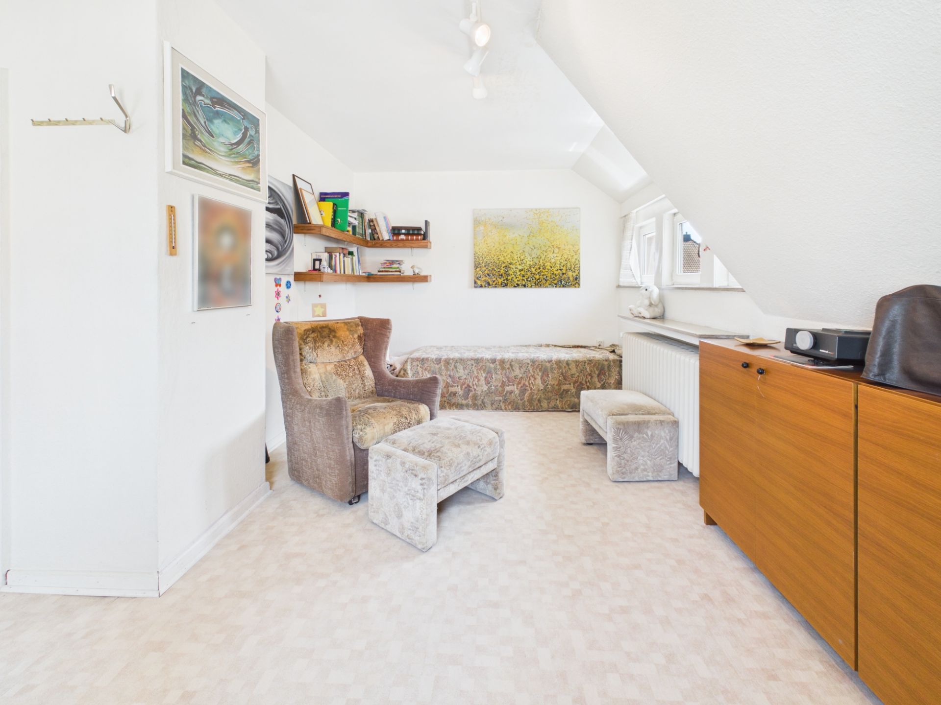 Helles Zimmer mit Sessel, Hocker, Regal, Kommode und einem Bett unter Dachschräge.