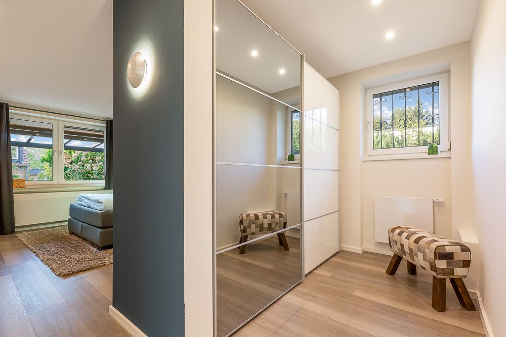 Modernes Schlafzimmer mit großem Spiegelkleiderschrank, zwei Fenster und zwei gepolsterten Bänken.