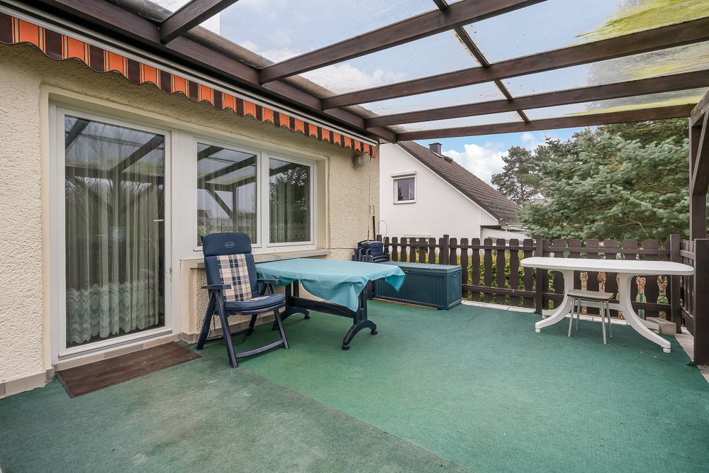 Überdachte Terrasse mit Tisch, zwei Stühlen und grünem Teppich, Blick auf Garten und Nachbarhaus.