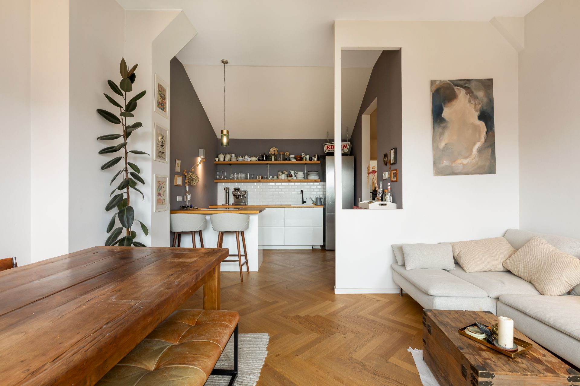 Helles Wohnzimmer mit Holztisch, Sofa, offener Küche und Zimmerpflanze.