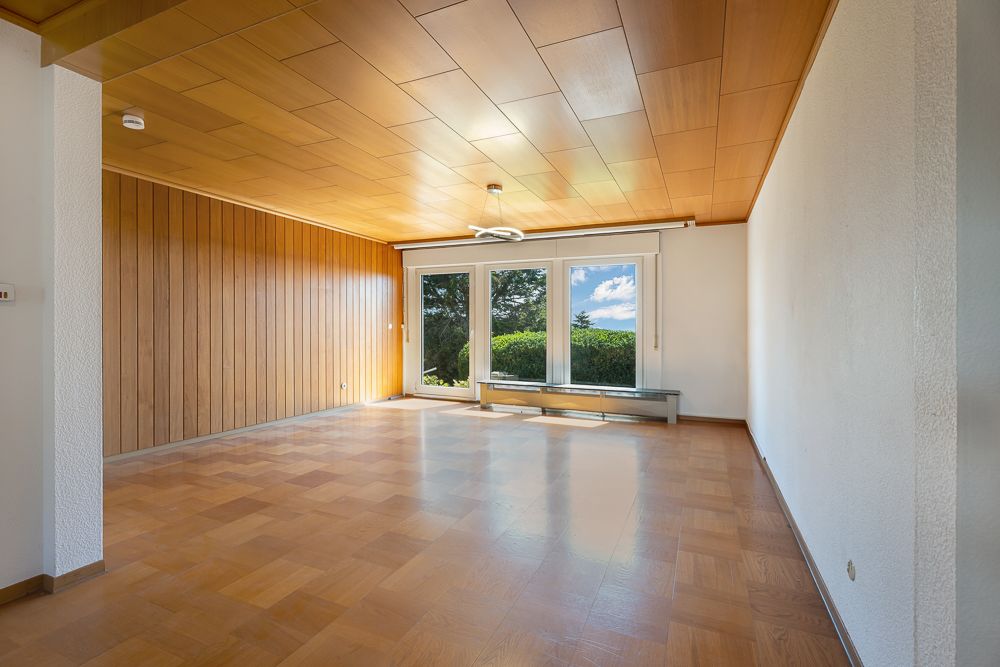 Leeres Wohnzimmer mit Holzdecke, Holzfußboden und großen Fenstern mit Blick ins Grüne.