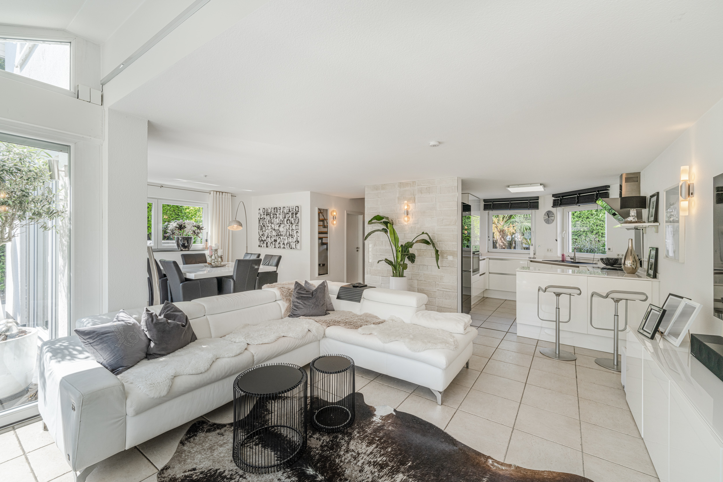 Helles Wohnzimmer mit weißem Sofa, offener Küche, Esstisch und großen Fenstern.