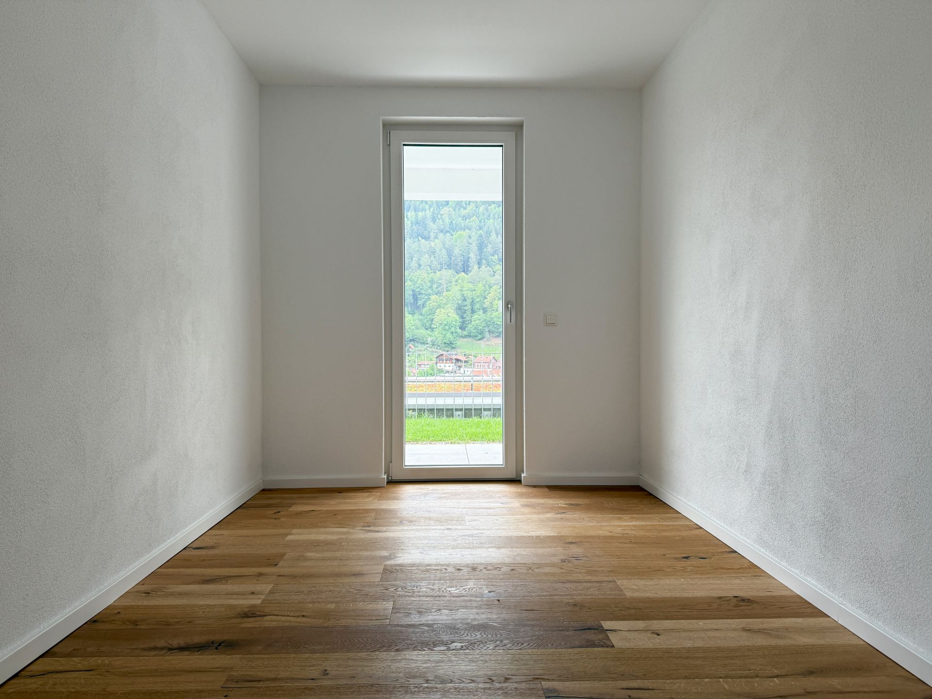 Leerzimmer mit Holzboden und Tür zum Balkon, Blick auf Wald und Häuser.