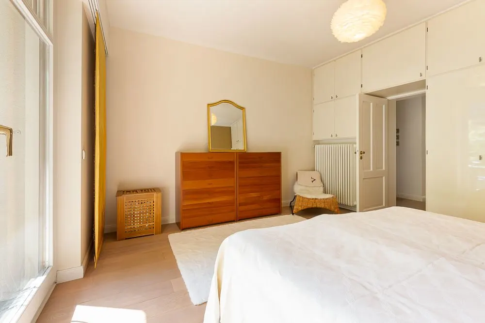 Helles Schlafzimmer mit Holzkommode, Spiegel, Stuhl, Teppich und Einbauschränken.