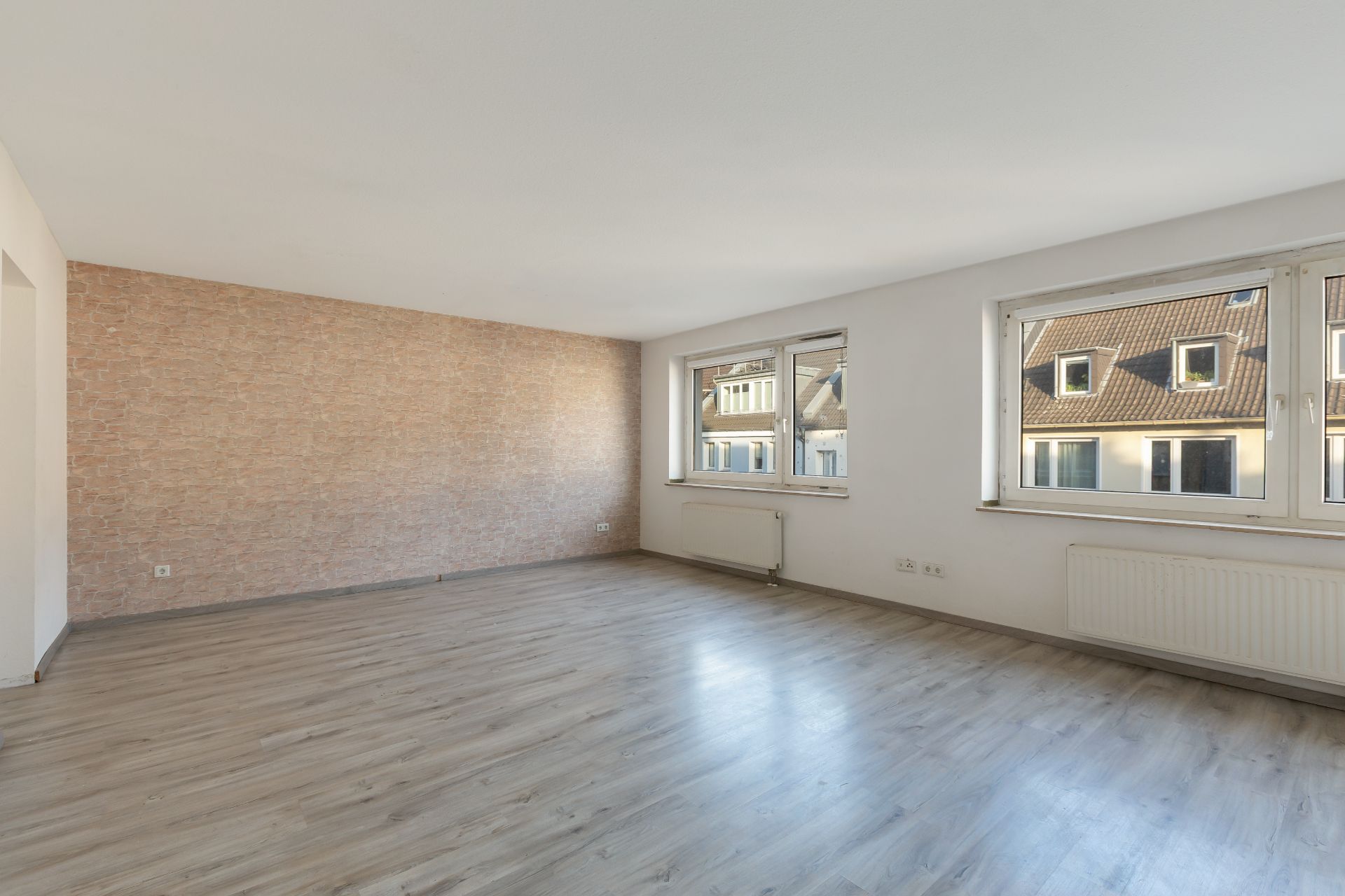 Helles, leeres Wohnzimmer mit Parkettboden, zwei Fenstern und gemusterter Wand.