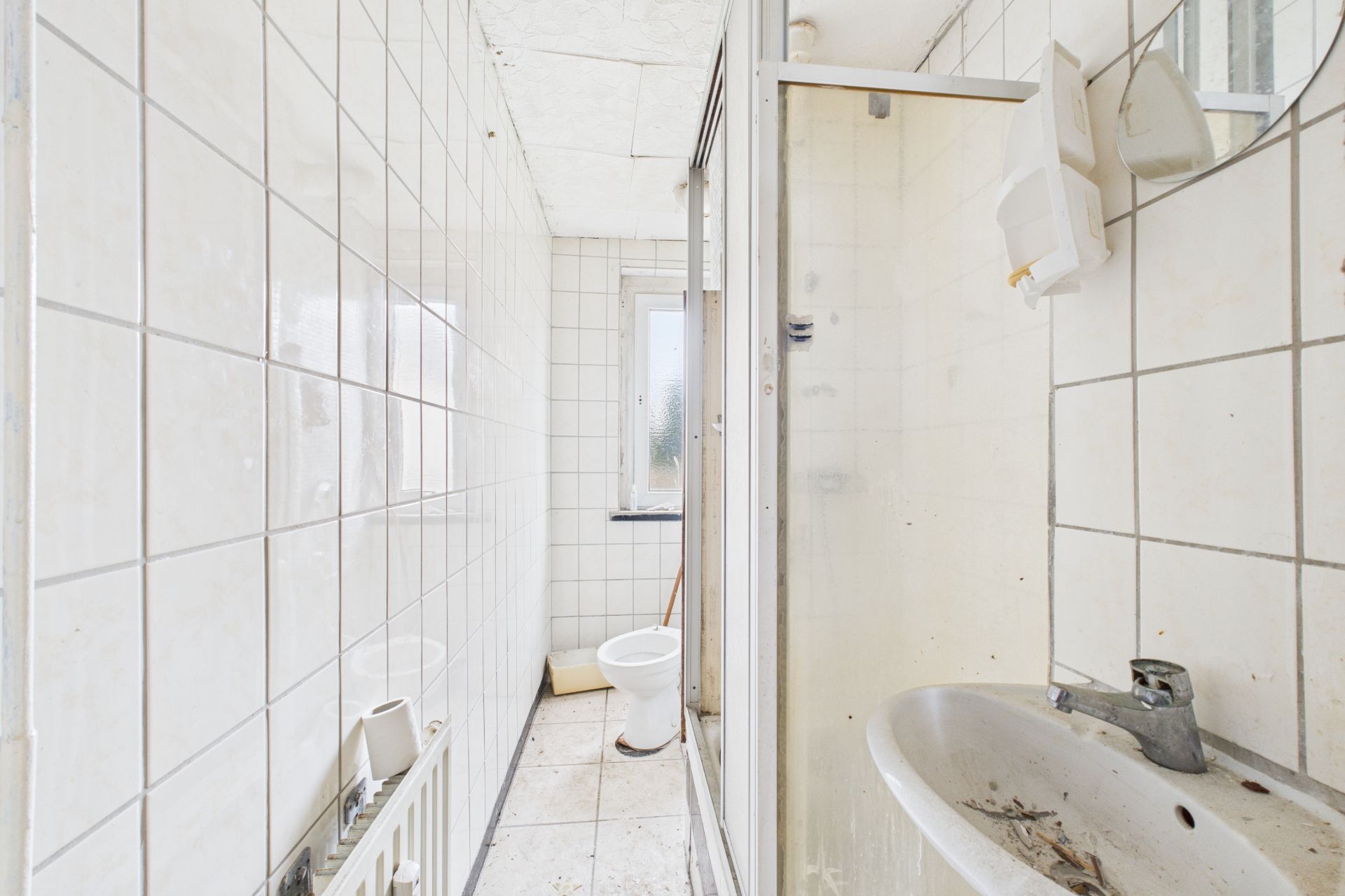 Schmales Badezimmer mit Dusche, Waschbecken, Toilette und Fenster, viele weiße Fliesen.