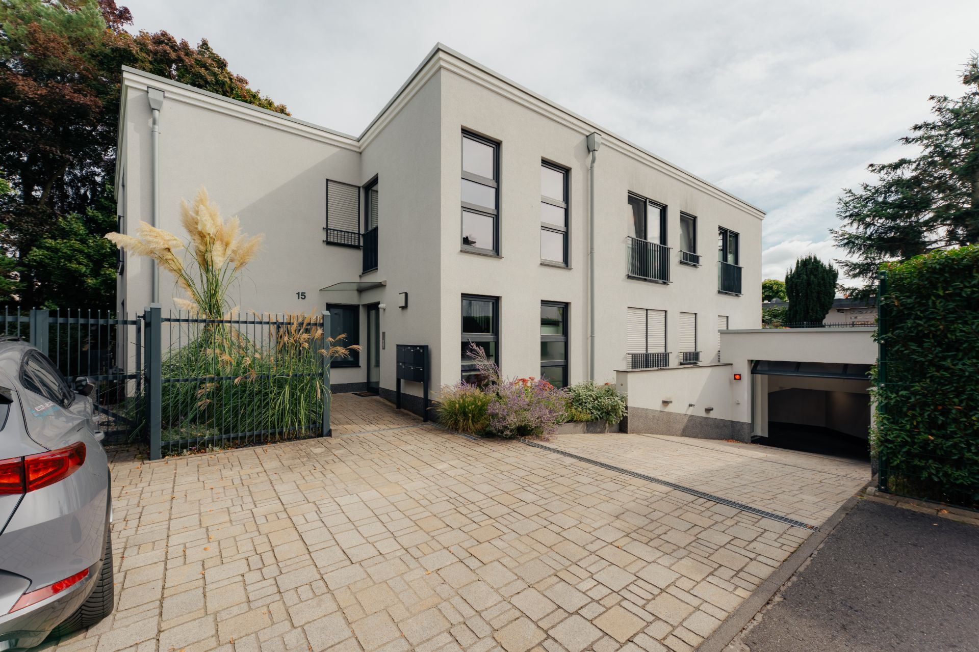 Moderne, weiße Wohnhausfassade mit großer Einfahrt, Pflanzen und einem geparkten Auto.