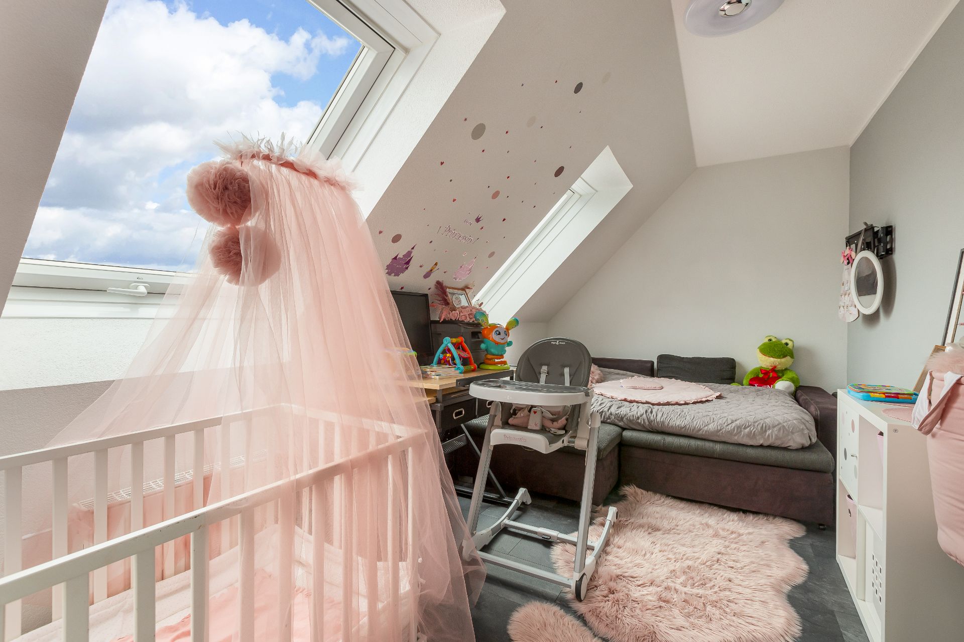 Kinderzimmer mit Babybett, Hochstuhl, Sofa, Dachfenstern und rosa Dekoration.