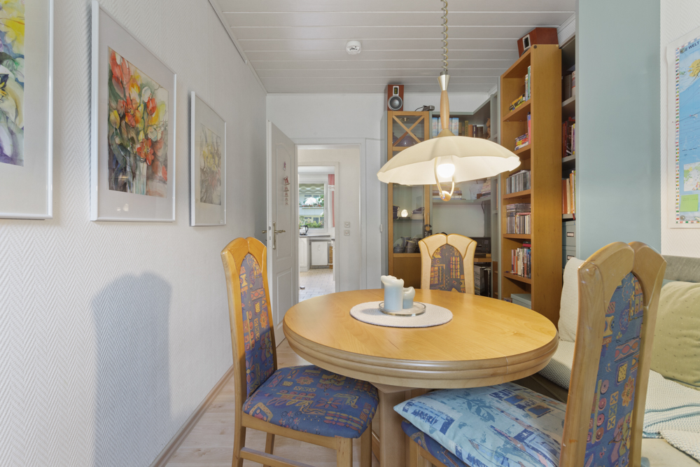 Essbereich mit rundem Holztisch, vier Stühlen, Wandbildern und Regal im Hintergrund.