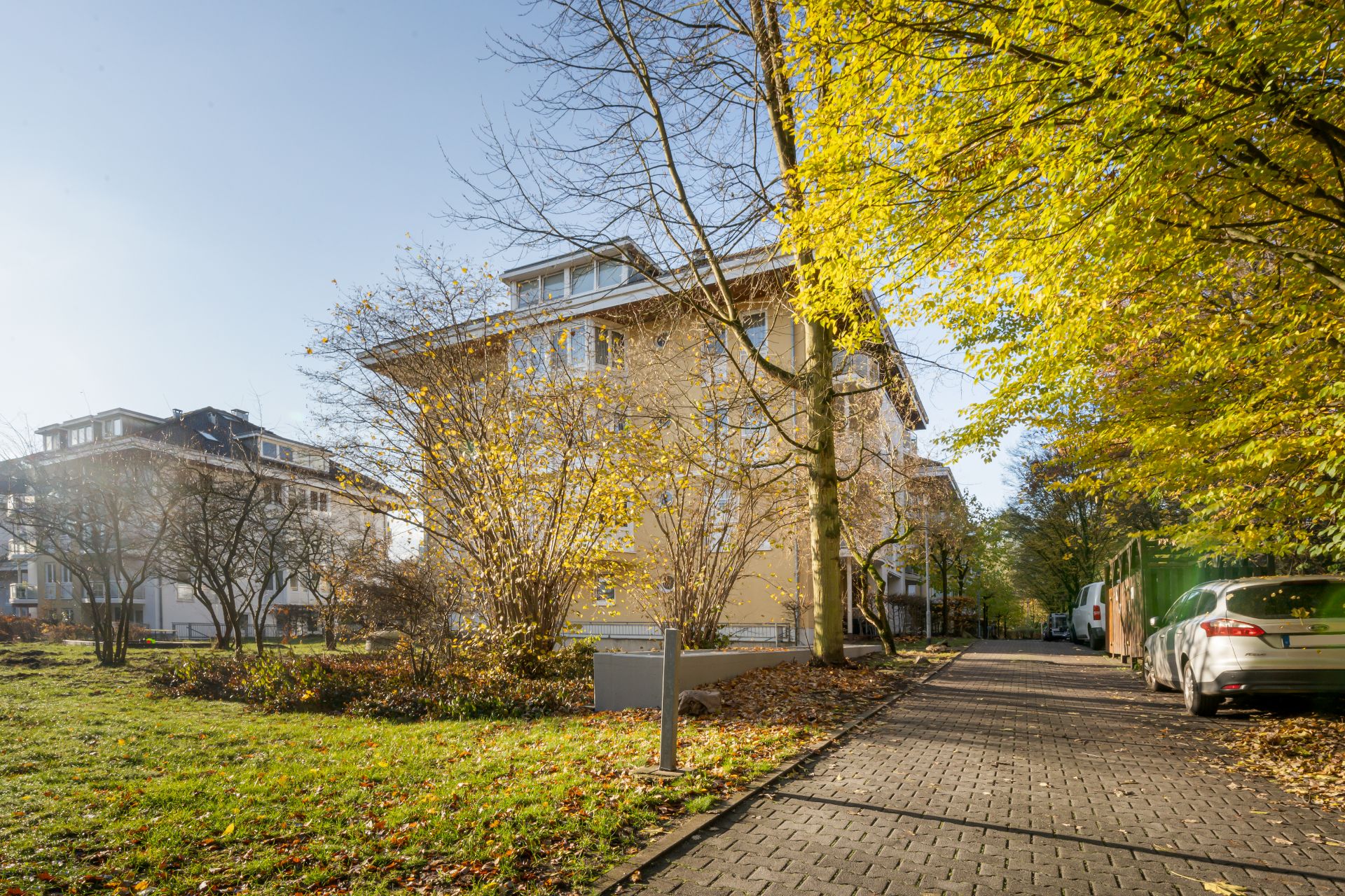 Mehrfamilienhaus mit gelber Fassade, Bäumen und parkenden Autos im herbstlichen Außenbereich.