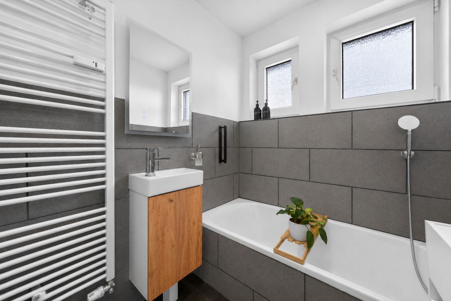 Modernes Badezimmer mit Badewanne, Waschbecken, Handtuchheizkörper und zwei Fenstern.