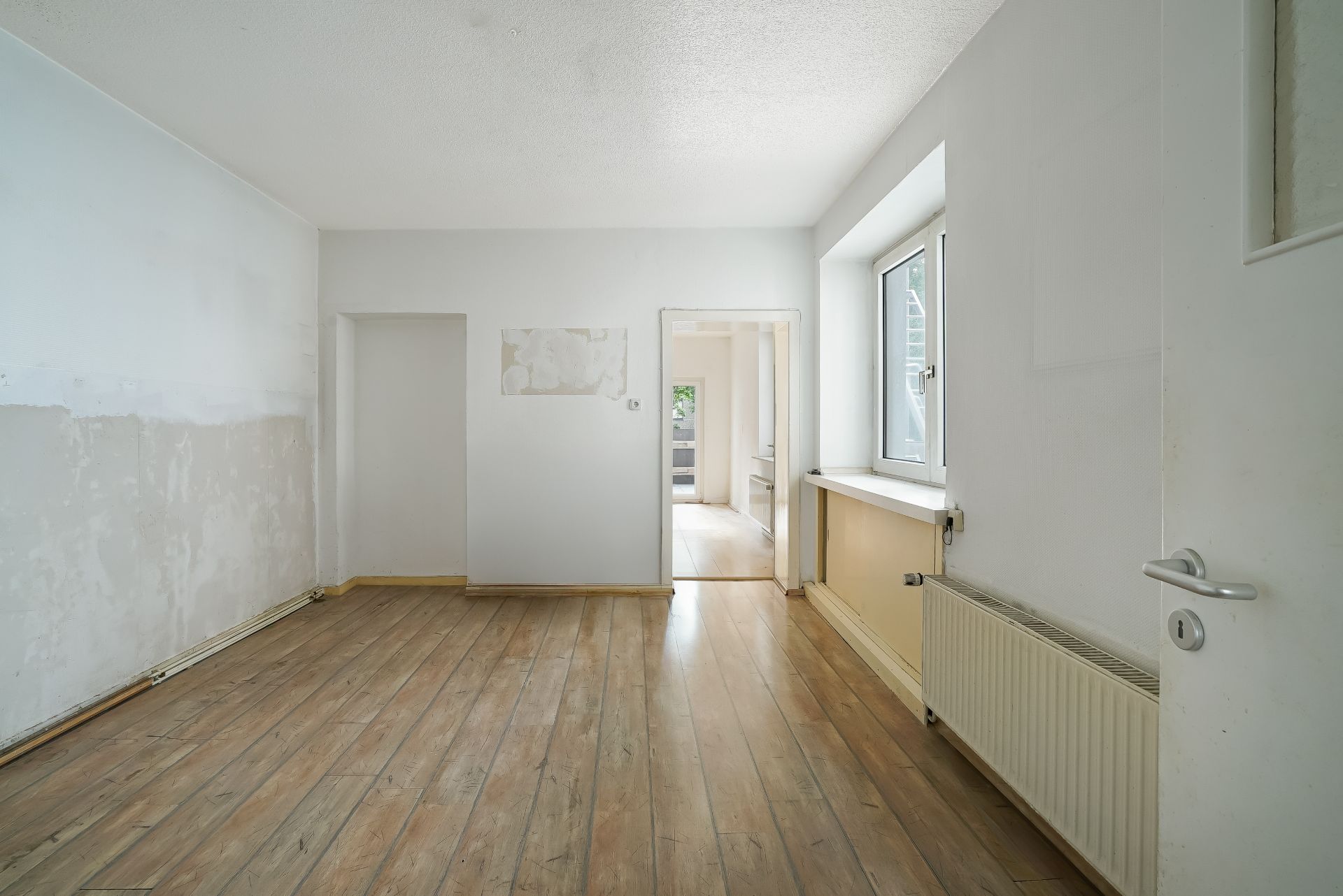 Leerer Raum mit Holzboden, weißer Wand, Fenster und Heizkörper.