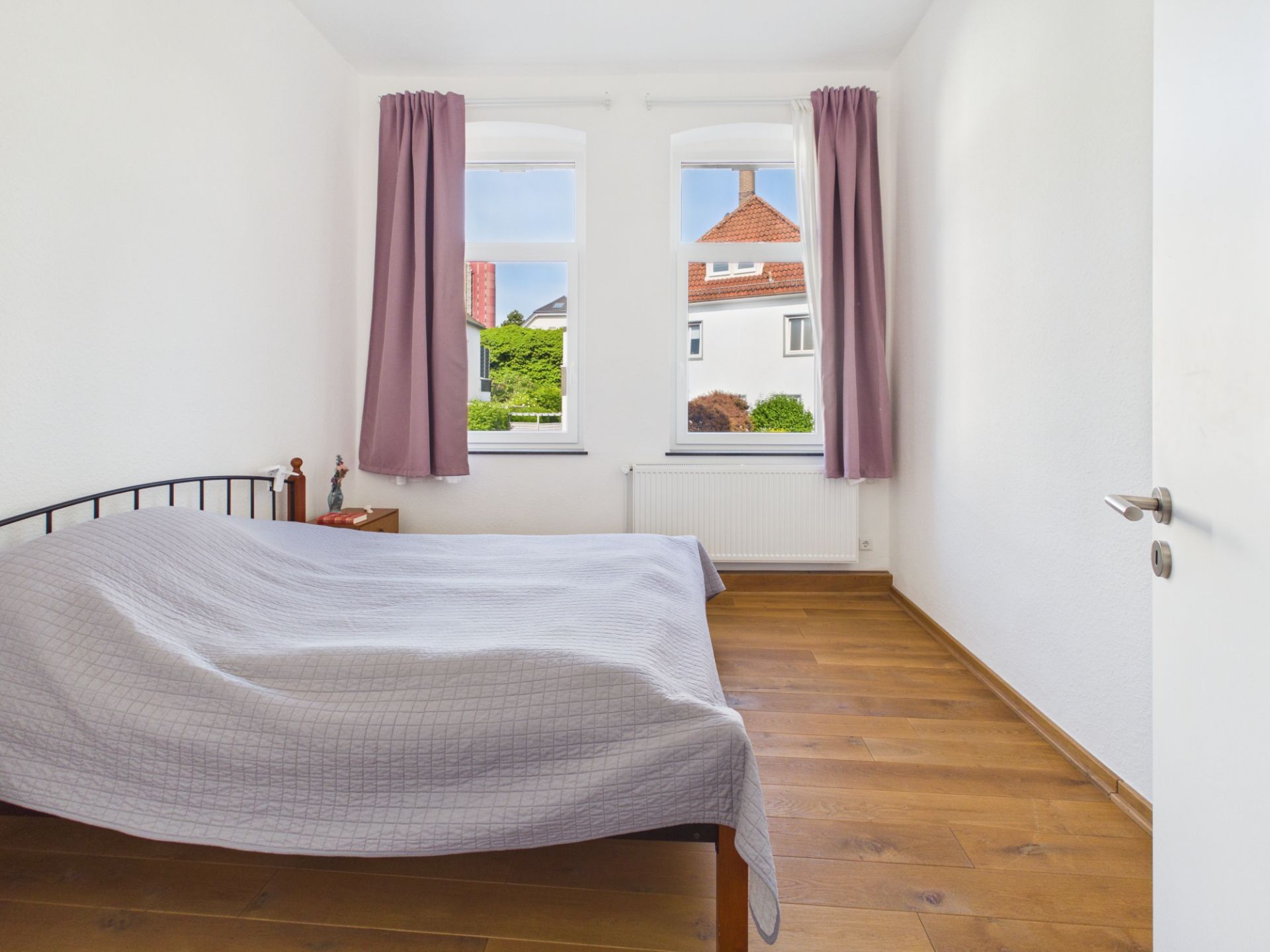 Schlafzimmer mit Holzboden, Doppelbett, zwei Fenstern und lila Vorhängen.