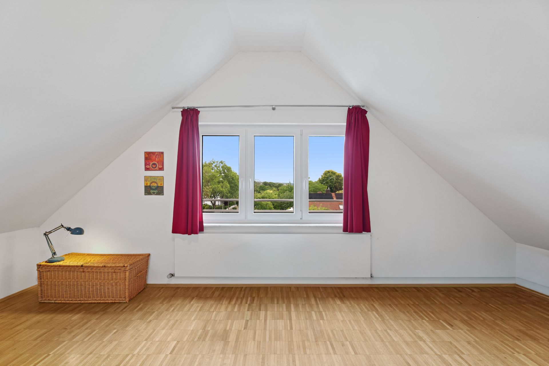 Helles Dachzimmer mit Holzfußboden, großem Fenster mit roten Vorhängen und Korbtruhe.