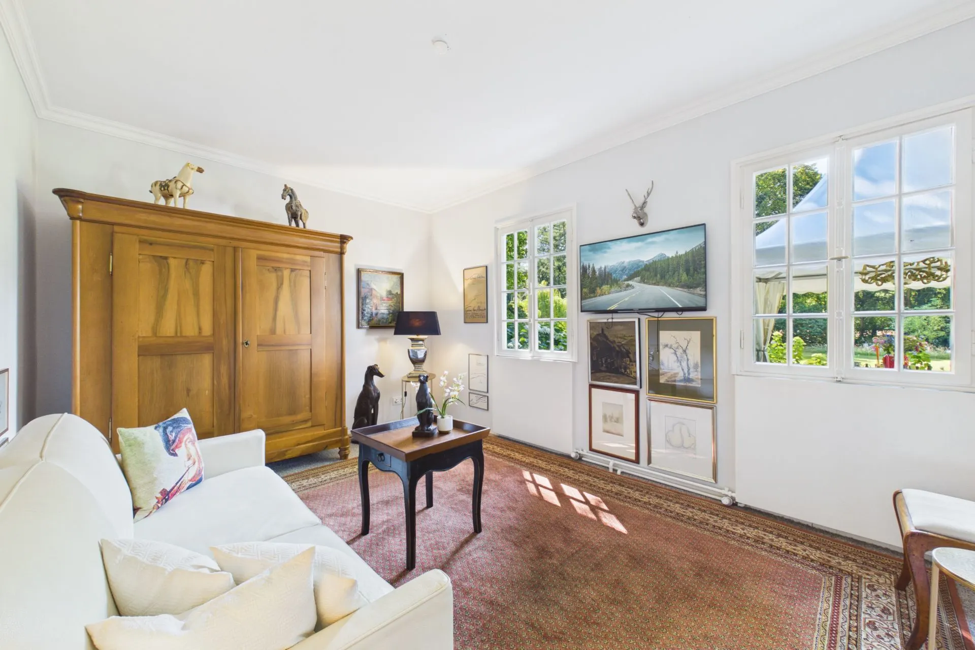 Helles Wohnzimmer mit Sofa, Holztisch, Teppich, großem Schrank und Fenstern mit Gartenblick.