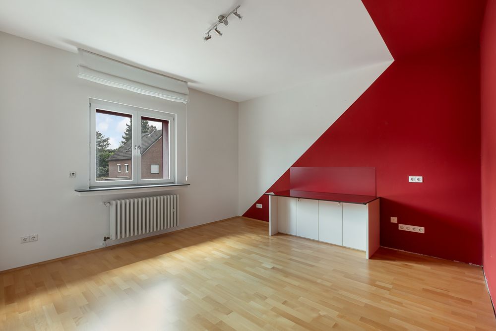 Helles Zimmer mit Holzboden, roter Akzentwand, Fenster und Einbauschrank.