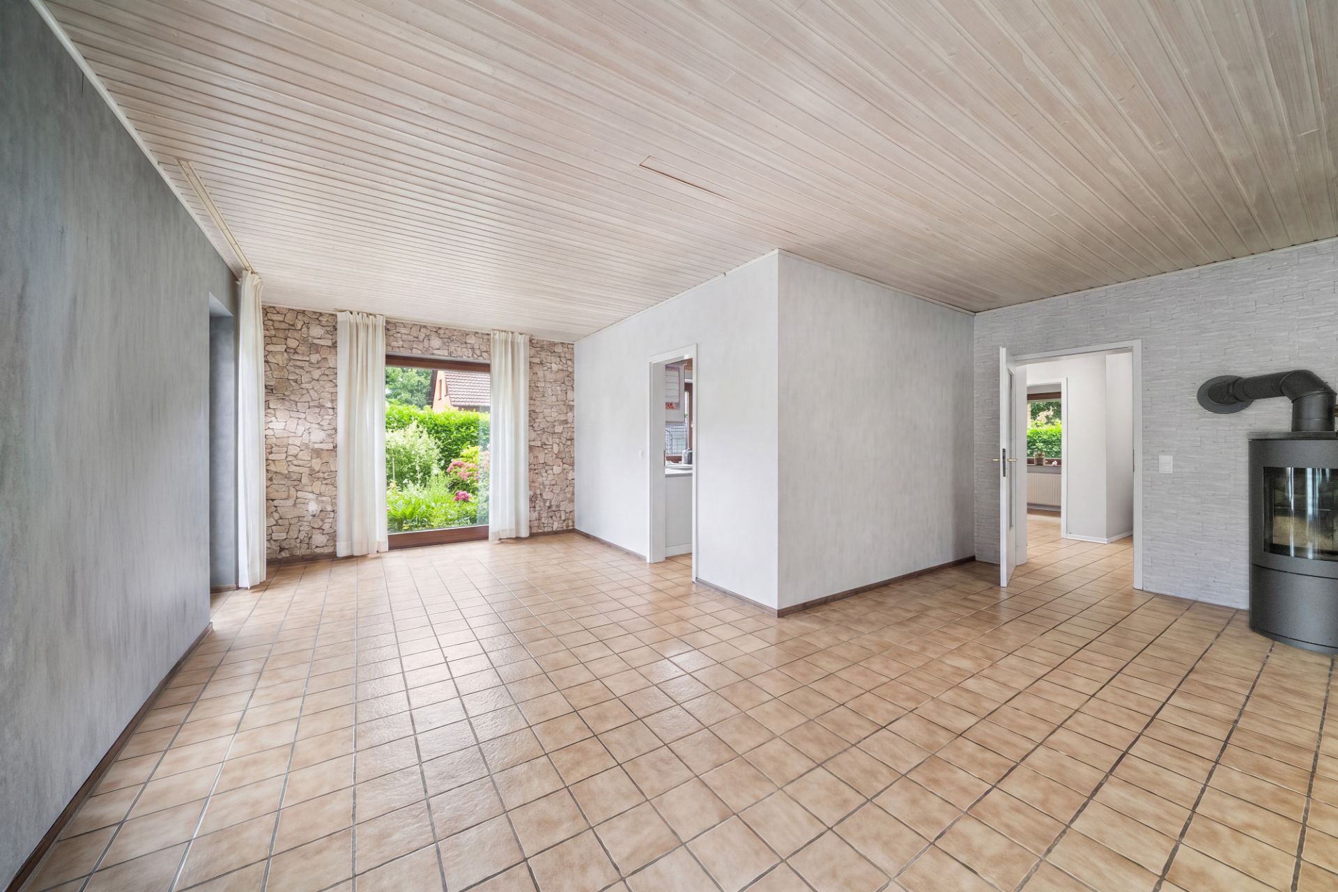 Helles Wohnzimmer mit Fliesenboden, großen Fenstern, weißen Wänden und Kaminofen.