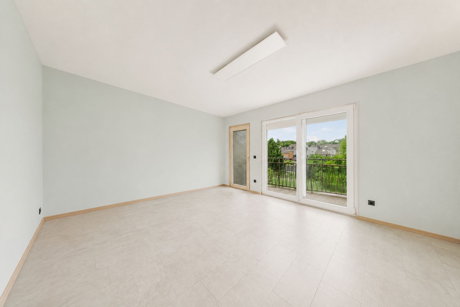 Leeres, helles Zimmer mit großen Fenstern und Blick auf einen Balkon mit grünen Bäumen.