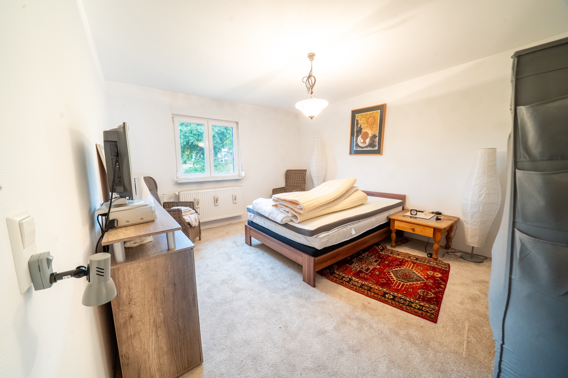 Helles Schlafzimmer mit Bett, Teppich, Kommode, Fernseher, Sessel und Fenster.