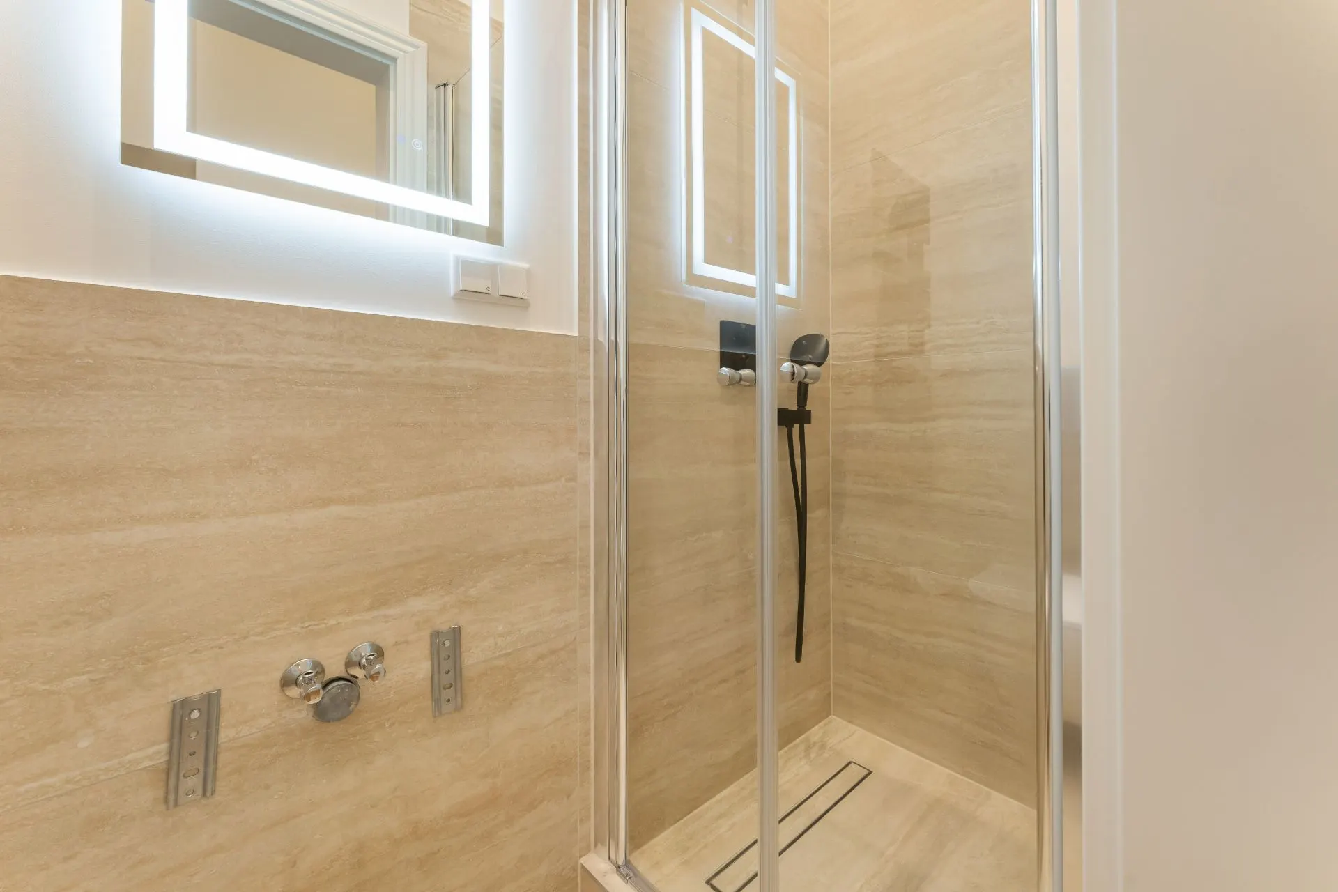 Moderne Dusche mit Glastür, Duschkopf und beleuchtetem Spiegel an hellen Fliesenwänden.