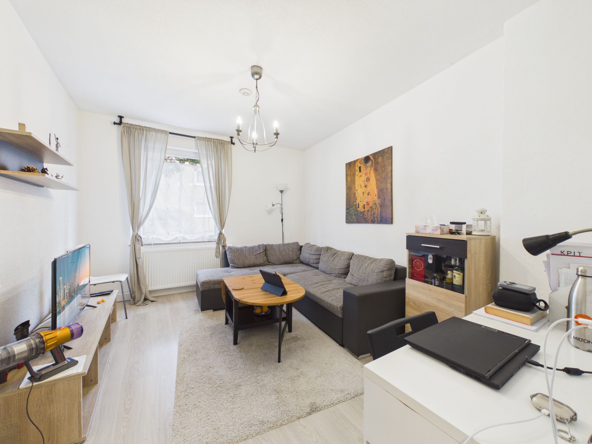 Helles Wohnzimmer mit grauem Sofa, Couchtisch, Fernseher, Schreibtisch und Fenster mit Vorhängen.