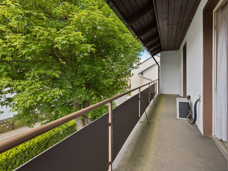 Langer Balkon mit Überdachung, Blick auf grünen Baum und Klimagerät an der Wand.
