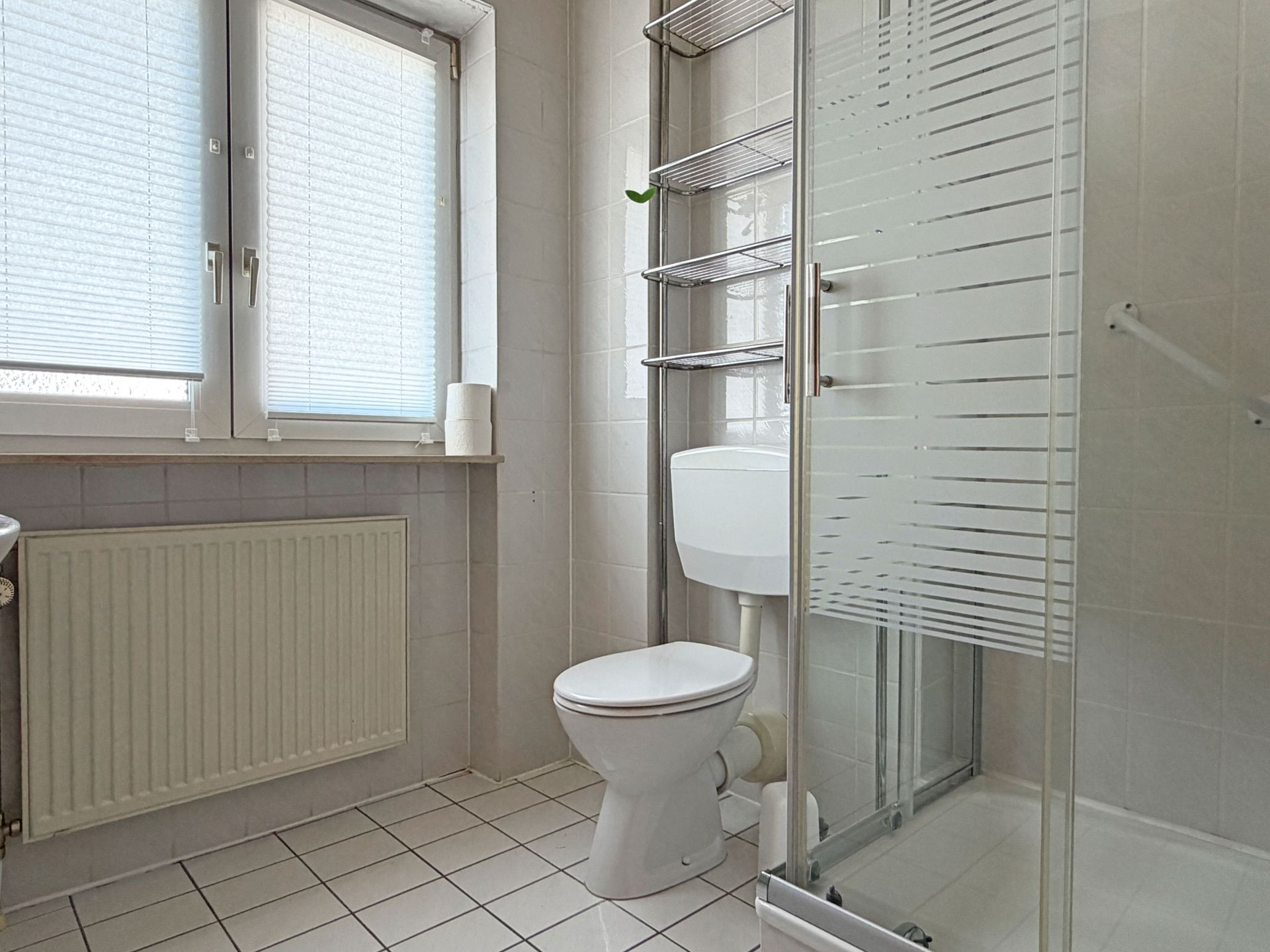 Helles Badezimmer mit Toilette, Dusche, Regal, Heizung und Fenster mit Jalousien.