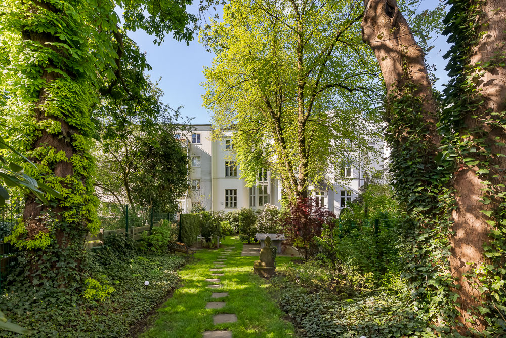 Grüner Garten mit Bäumen, Rasen und Sitzbank vor einem weißen Wohngebäude.