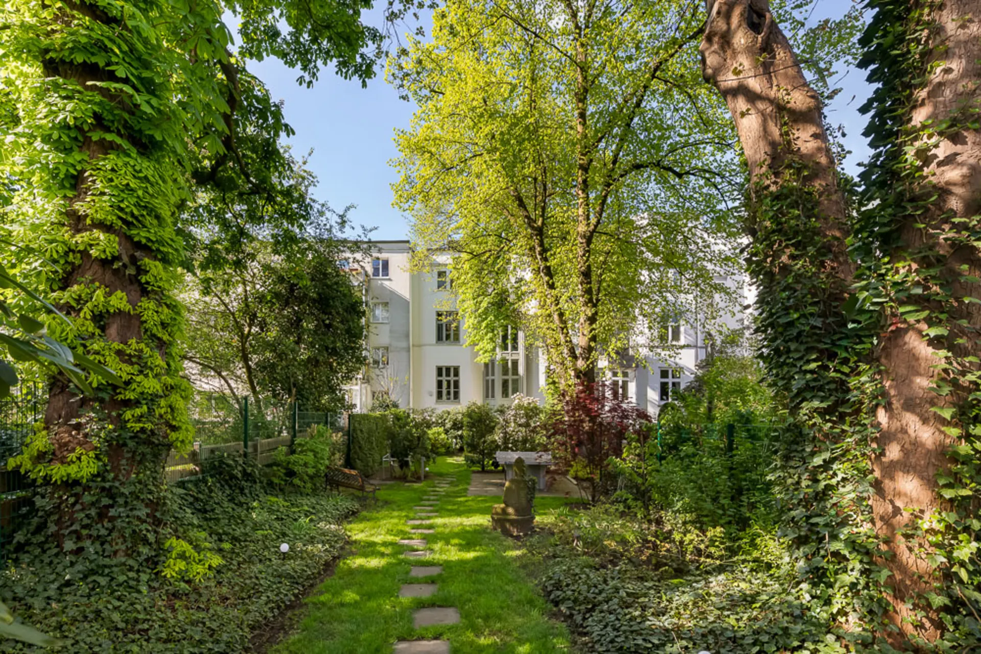 Grüner Garten mit Bäumen, Rasen und Sitzbank vor einem weißen Wohngebäude.