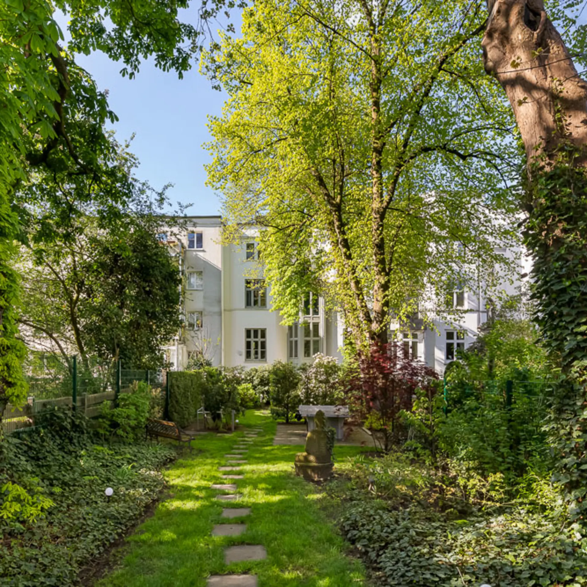 Grüner Garten mit Bäumen, Rasen und Sitzbank vor einem weißen Wohngebäude.