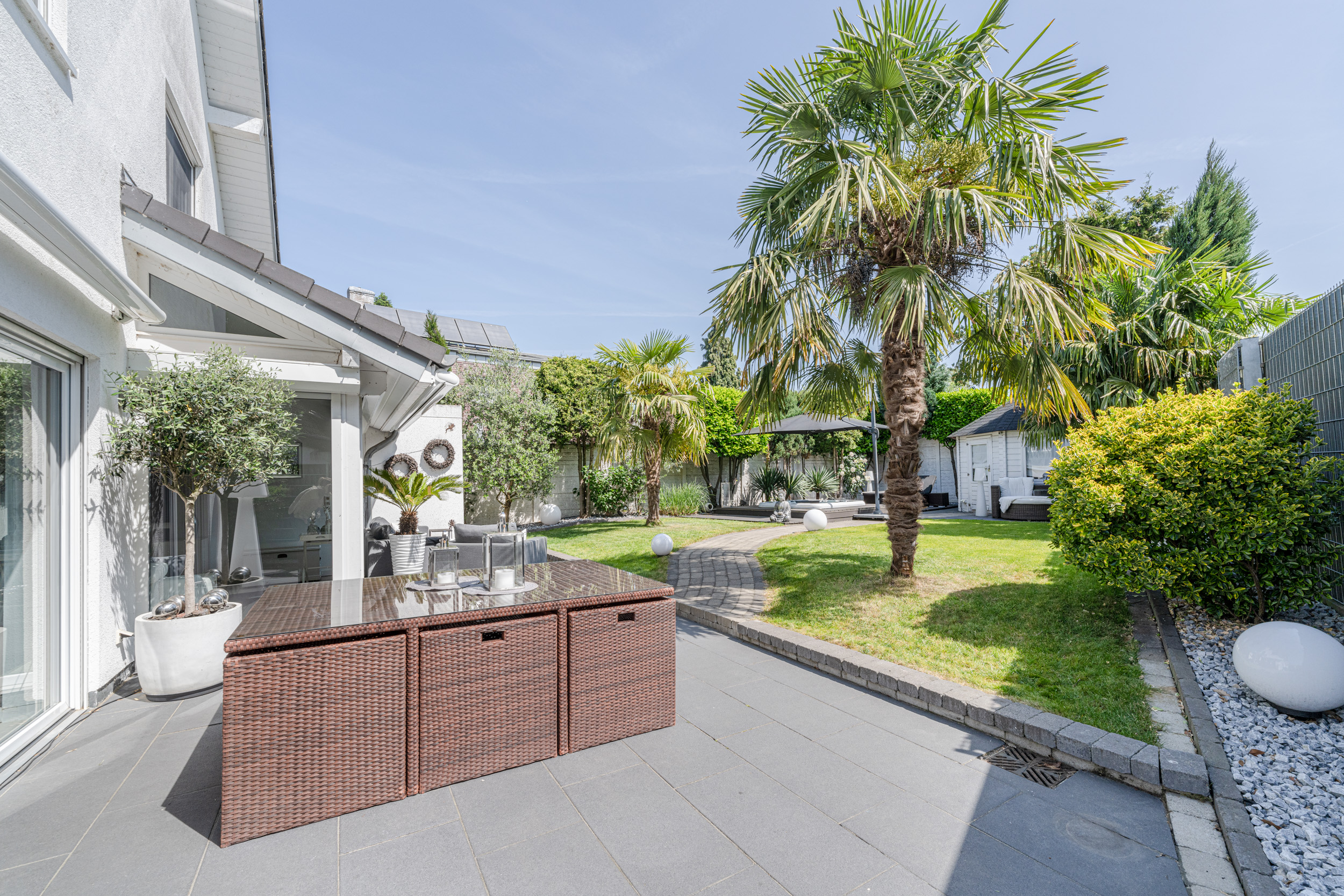 Garten mit Palmen, Terrasse mit Tisch und Stühlen, Rasenfläche und Gartenhaus bei sonnigem Wetter.