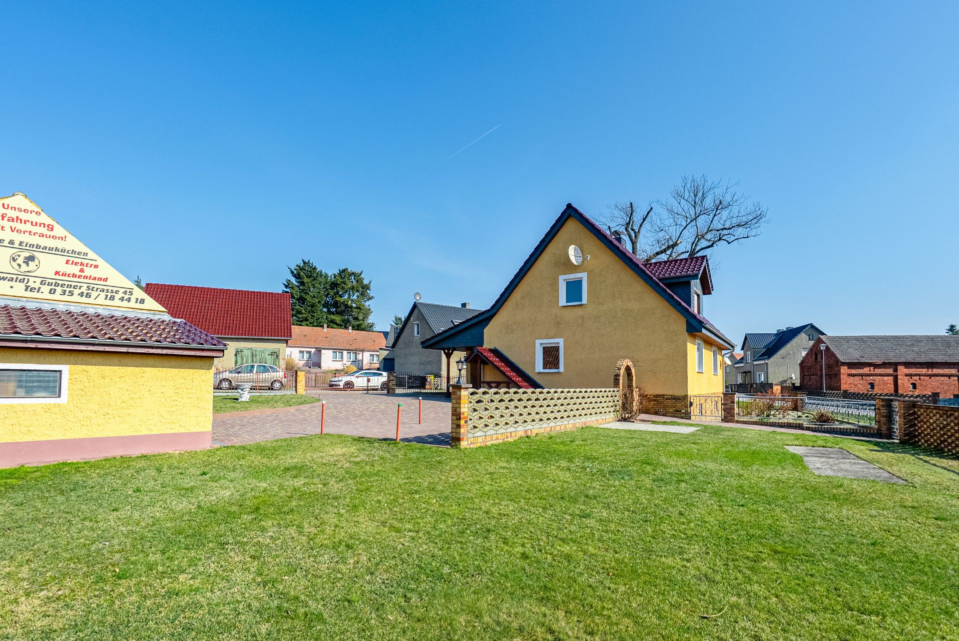Gelbes Einfamilienhaus mit Garten, gepflastertem Hof und Nebengebäude an einem sonnigen Tag.