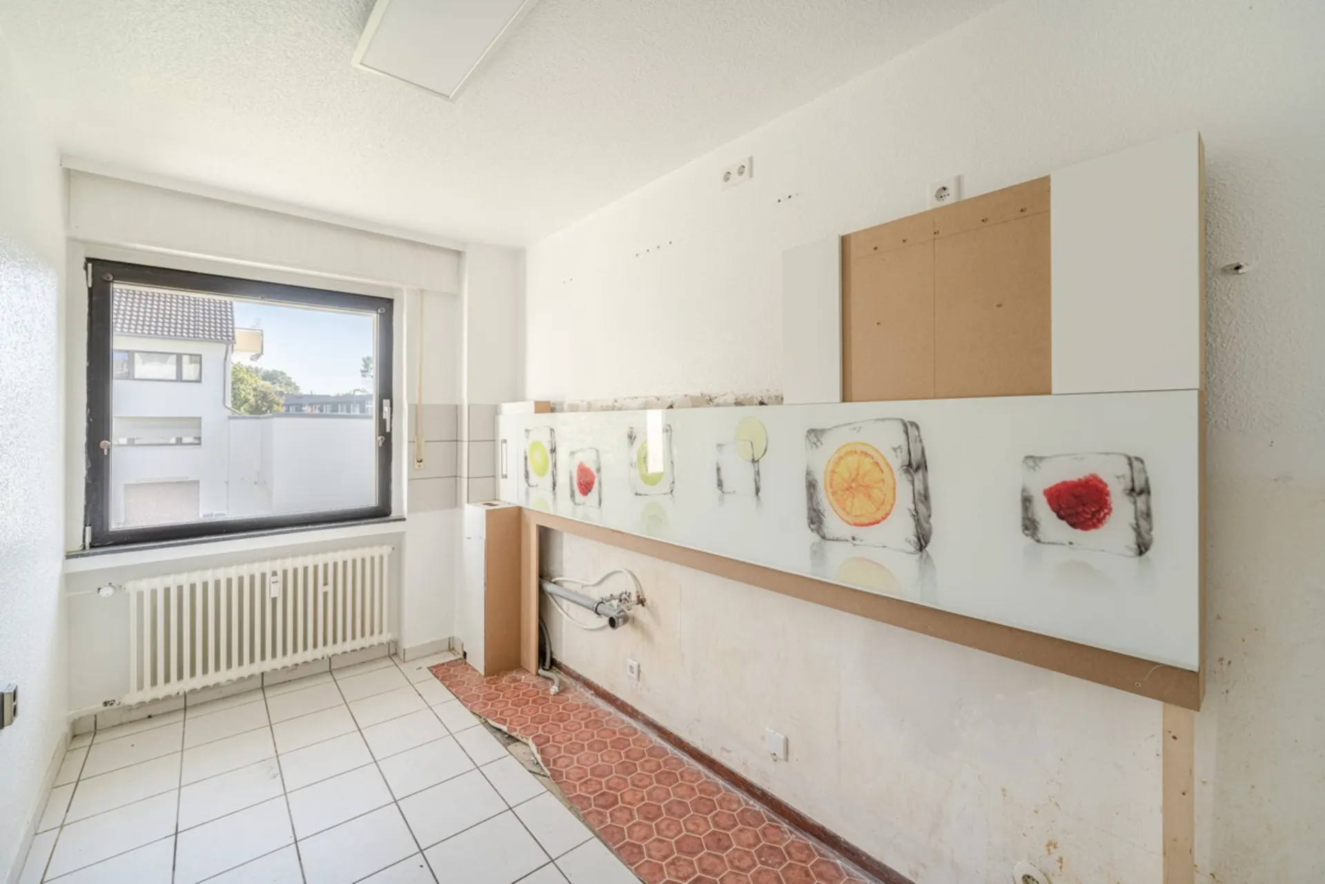 Leerer Küchenraum mit großem Fenster, Heizkörper und bunter Glasverkleidung mit Fruchtmotiven.