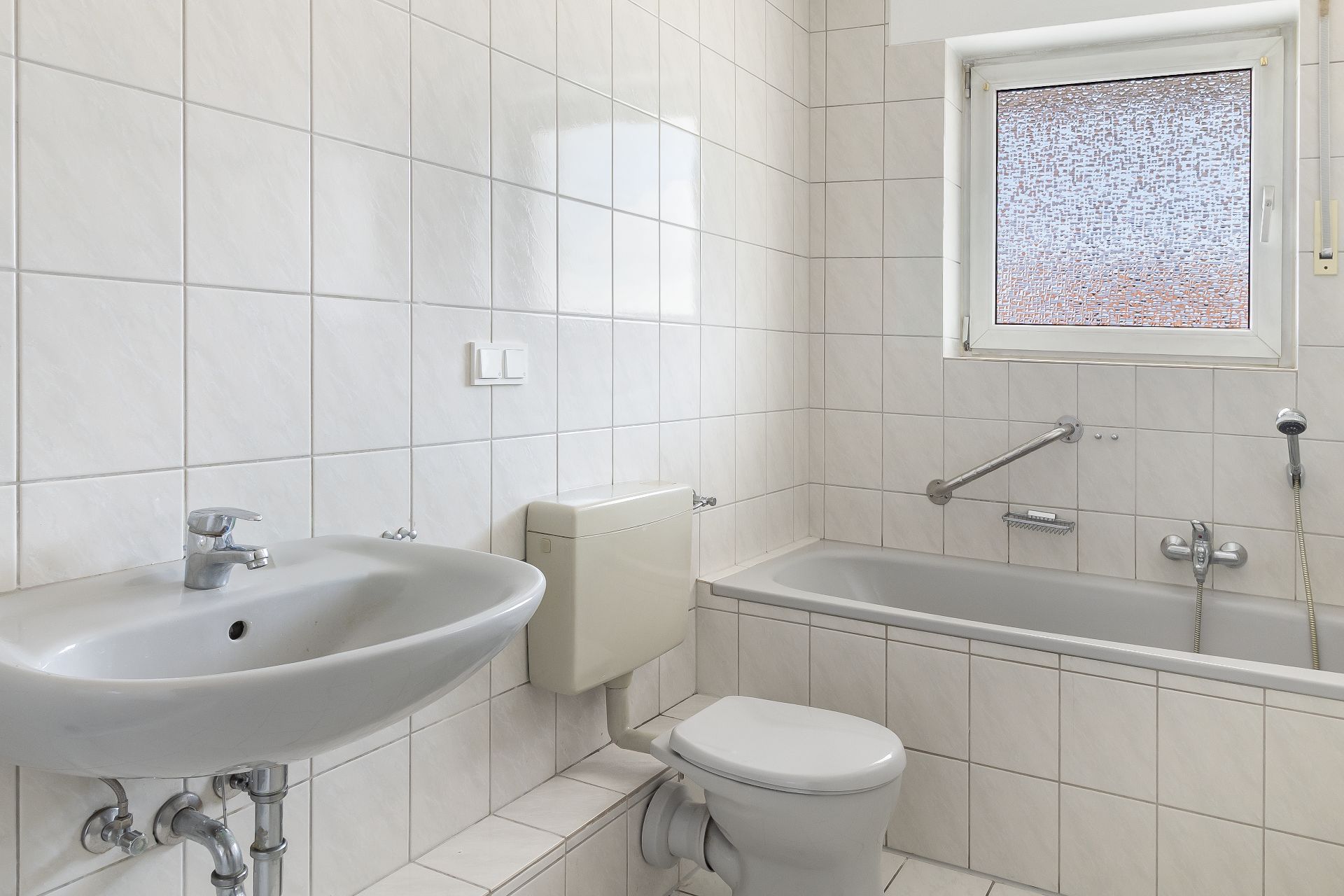 Helles Badezimmer mit Waschbecken, Toilette, Badewanne und Fenster mit Milchglas.