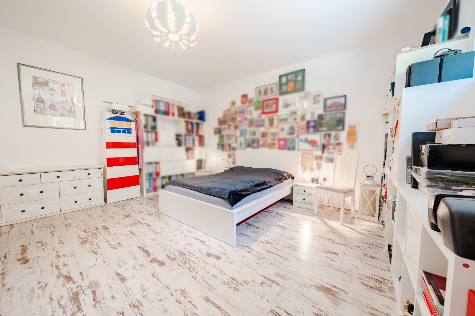 Helles Schlafzimmer mit weißem Bett, Bücherregalen, Kommode und vielen Bildern an der Wand.