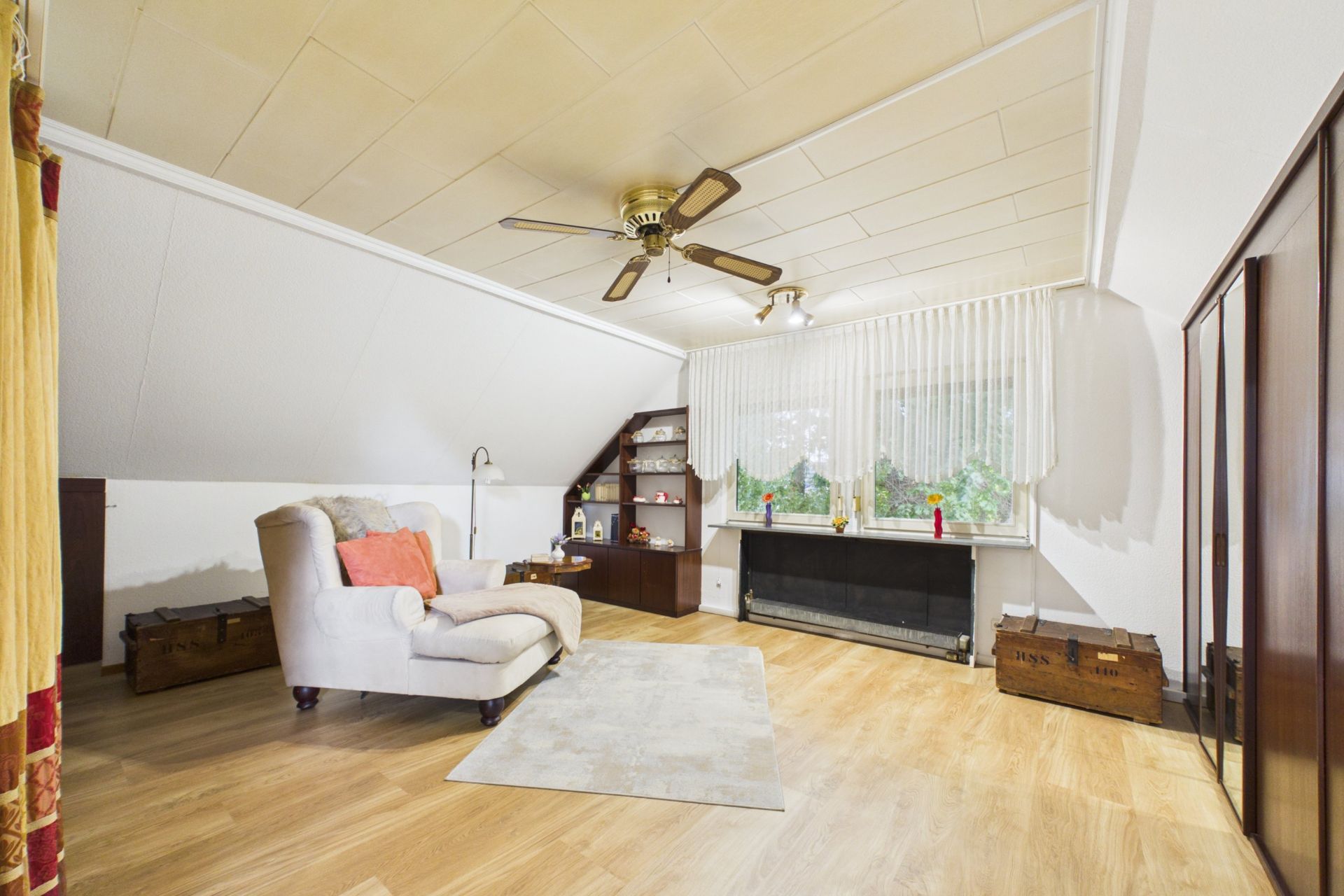 Helles Wohnzimmer mit Sessel, Teppich, Regal, großen Fenstern und Deckenventilator.