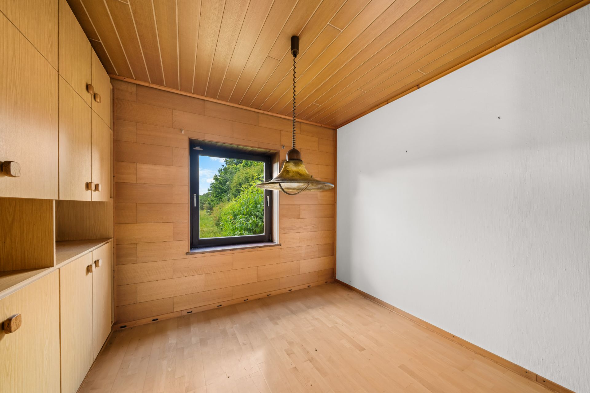 Leerer Raum mit Holzdecke, Holzschränken, einem Fenster und einer Hängelampe.