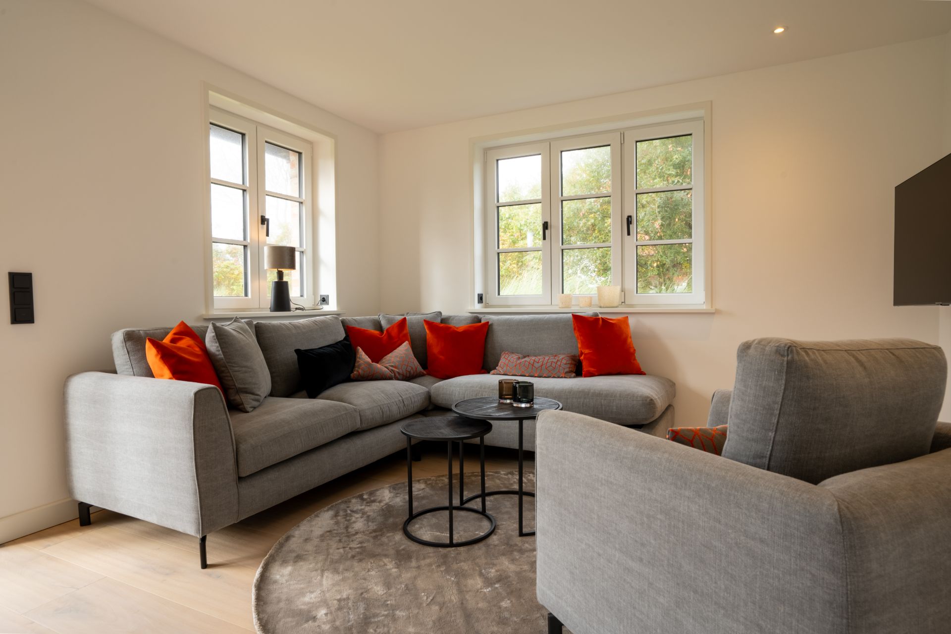 Wohnzimmer mit grauem Sofa, Sessel, roten Kissen, runden Tischen und großen Fenstern.