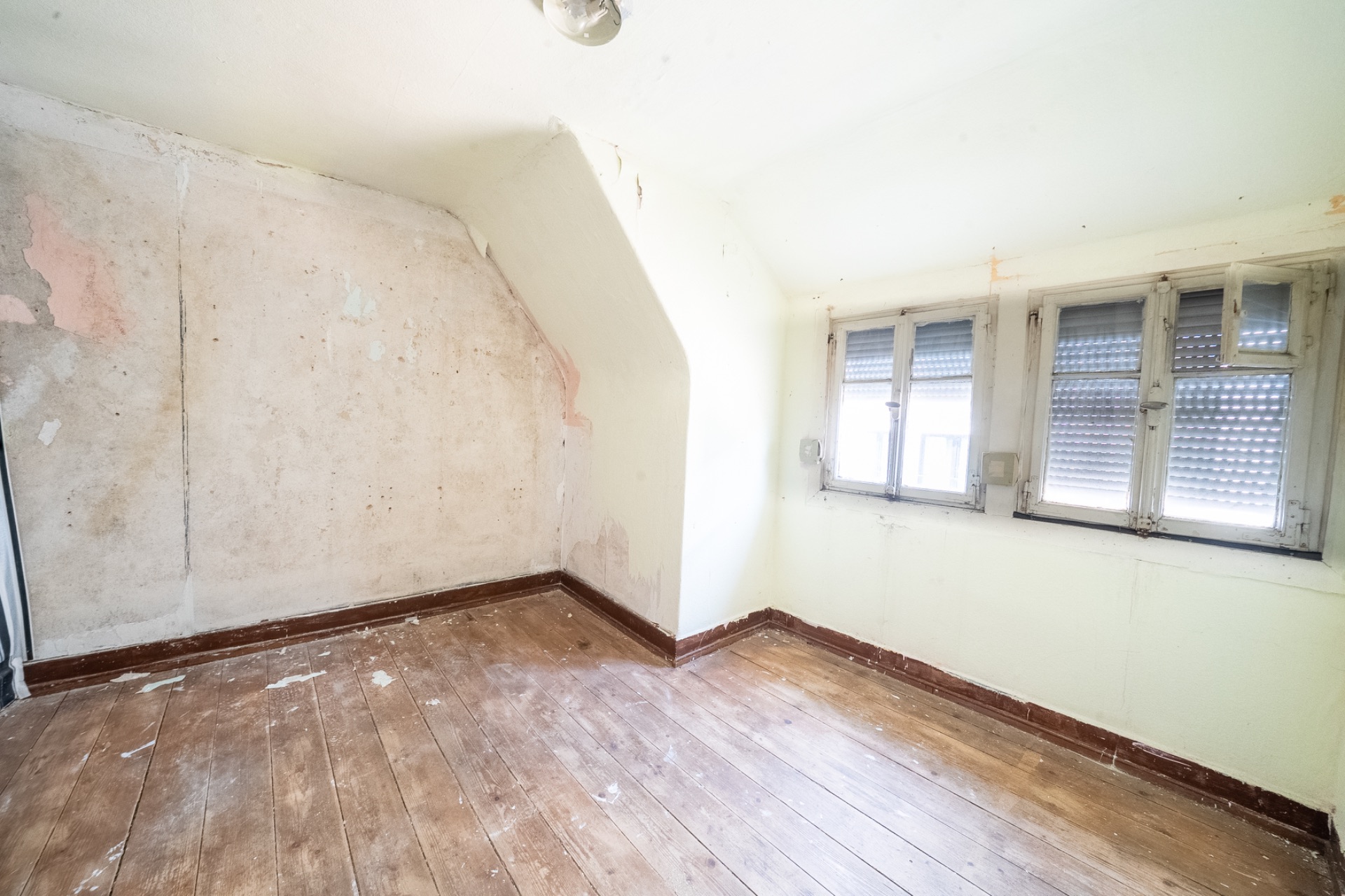 Leeres Zimmer mit Holzfußboden, schräger Wand und drei Fenstern mit Rollläden.