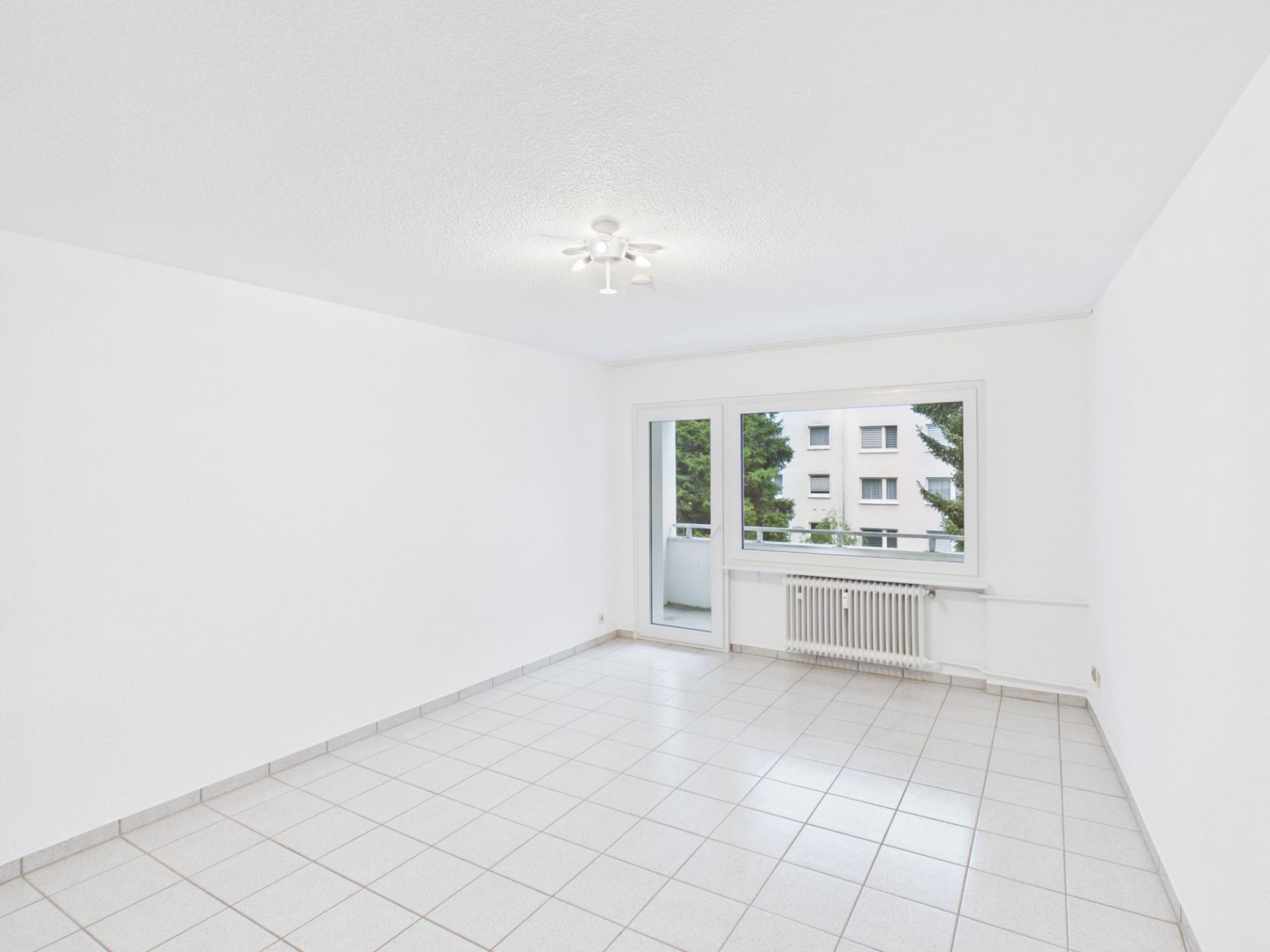 Heller, leerer Raum mit weißen Wänden, Fliesenboden, Fenster und Tür zum Balkon.