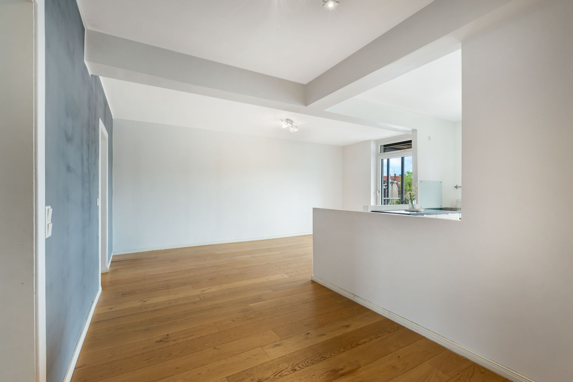 Helles Wohnzimmer mit Parkettboden und offener Küche, Fenster mit Blick nach draußen.