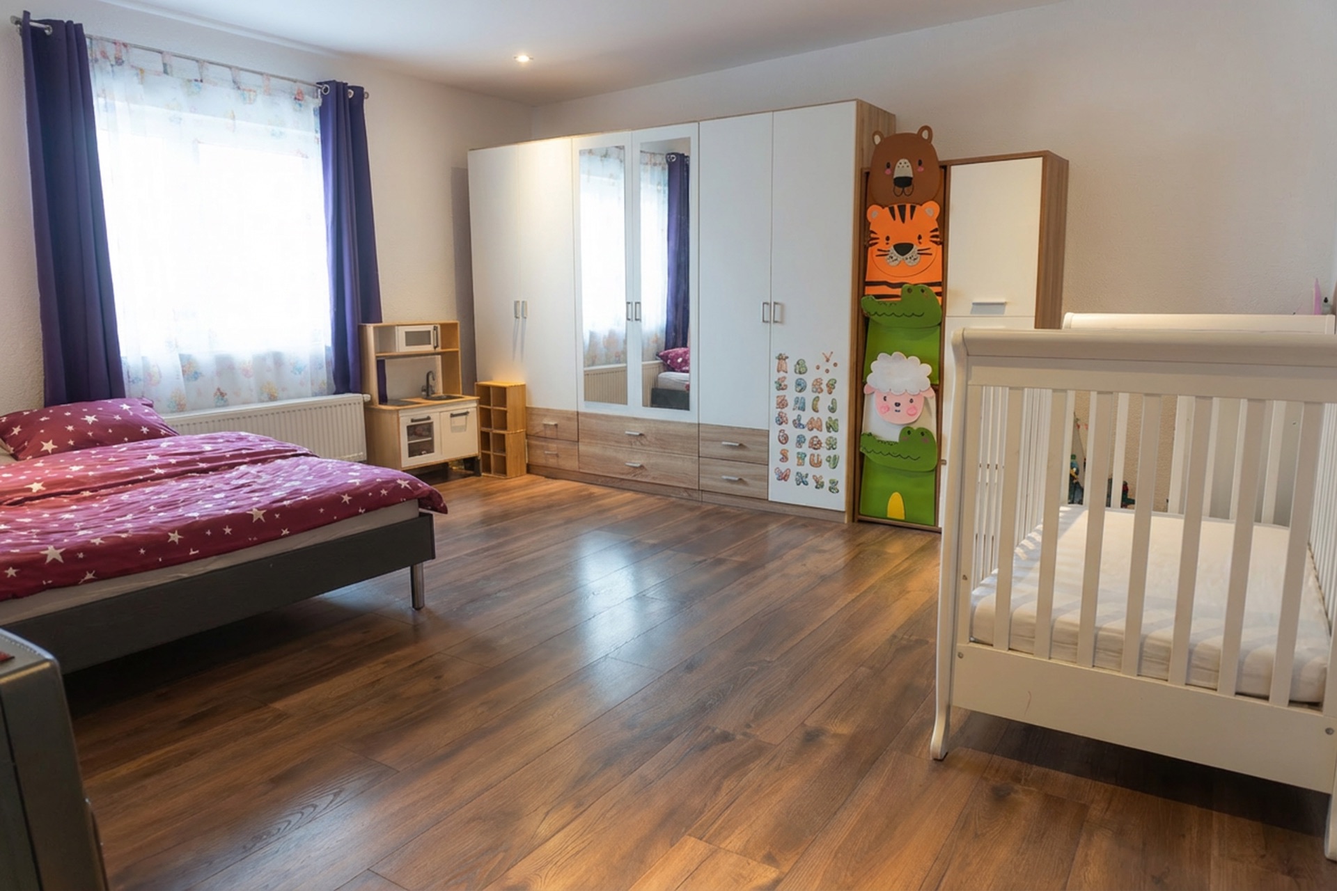 Helles Kinderzimmer mit Bett, Babybett, Kleiderschrank und Spielküche auf Holzfußboden.