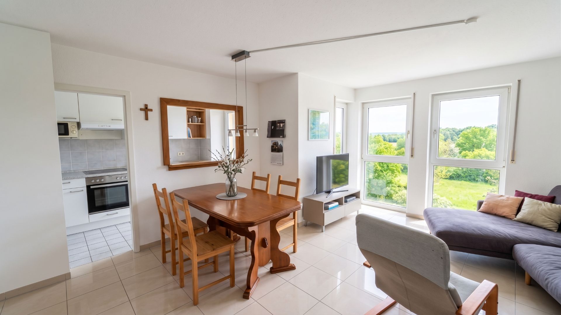 Helles Wohnzimmer mit Esstisch, Stühlen, Sofa, Sessel, Fernseher und großen Fenstern mit Blick ins Grüne.