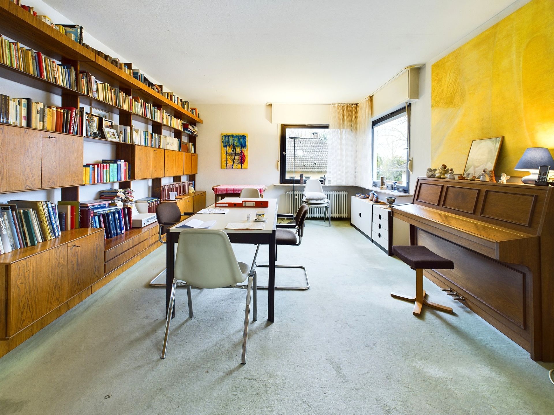 Helles Wohnzimmer mit Bücherregal, Tisch, Stühlen, Klavier und großen Fenstern.